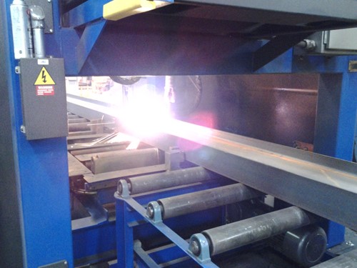 A steel beam being processed through an industrial machine with bright light indicating active cutting or welding, supported by rolling mechanisms.