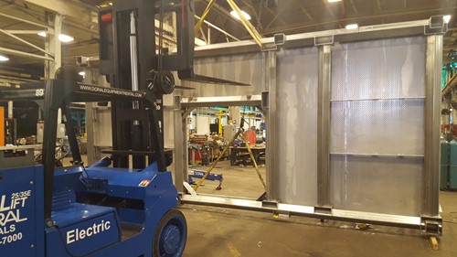 A blue electric forklift labeled “Versa-Lift Doral” assists in handling a large stainless steel platform inside an industrial workshop, with an overhead crane and additional equipment visible in the background.