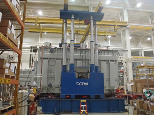 A heavy-duty Doral 600-ton gantry system is being used to lift a massive 300,000-pound transformer inside a spacious industrial facility, surrounded by equipment and shelving.