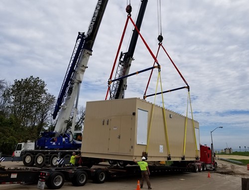 A large 90,000-pound electrical house is being lifted by two cranes using rigging straps, as workers supervise the operation on a construction site with a flatbed truck in the background.