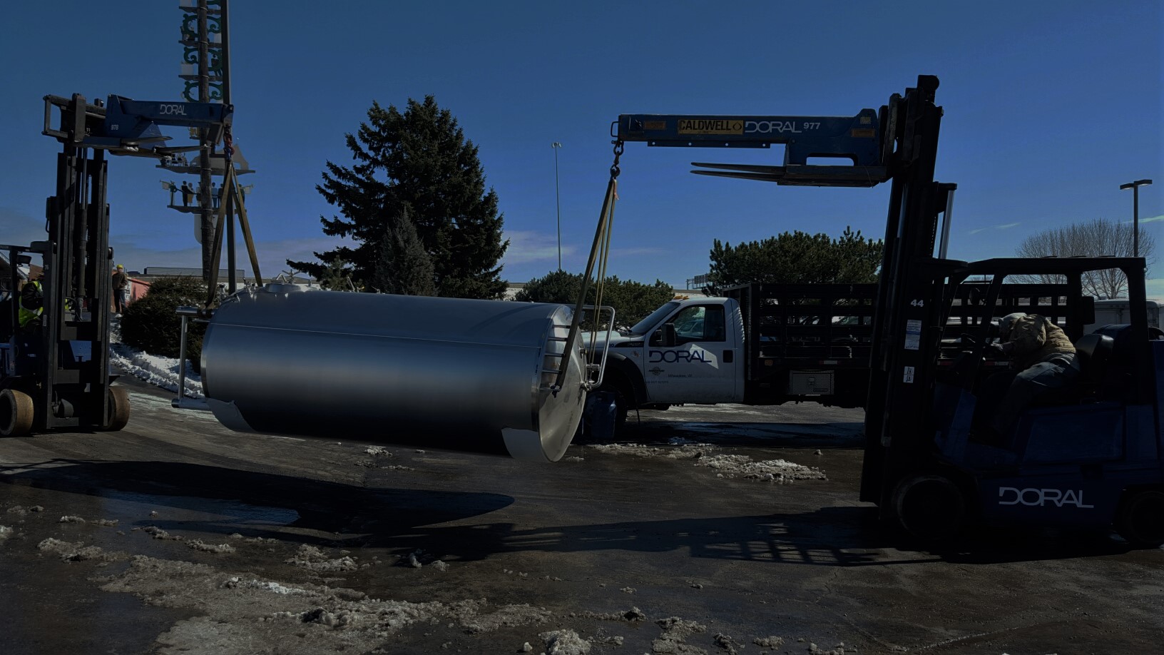 Two Doral forklifts are carefully maneuvering a large stainless steel tank suspended by rigging straps in an outdoor worksite on a clear day, with snow on the ground and a Doral service truck in the background.