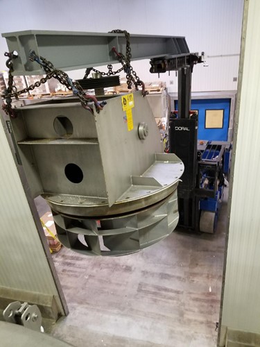A large spiral oven component is being maneuvered through a doorway using a blue Doral forklift and rigging chains in an industrial assembly operation.