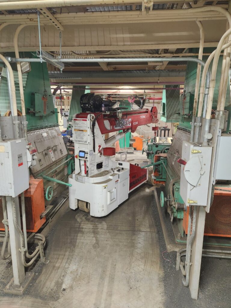 A compact Brandon Bailey crane is positioned in a wastewater plant, surrounded by industrial equipment, pipes, and machinery. The area is illuminated with overhead lights, highlighting the complex layout.