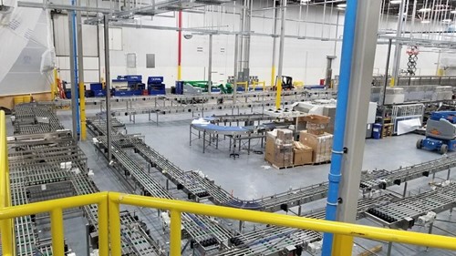 A large factory floor with multiple conveyor systems being installed, featuring interconnected tracks, equipment, and boxes of materials, viewed from an elevated perspective.