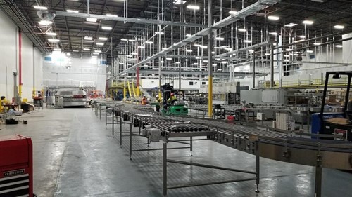 A factory floor showcasing a newly installed conveyor system with interconnected metal tracks extending across the space, surrounded by equipment, workers, and industrial lighting overhead.