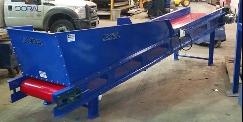 A custom-built blue conveyor system by Doral, featuring a red roller and designed for industrial material handling, placed in a workshop with vehicles in the background.