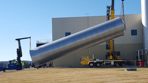A large stainless steel water silo is being erected using a crane and rigging equipment, with a forklift assisting on one side. The operation is taking place outside a modern industrial facility on a clear day.