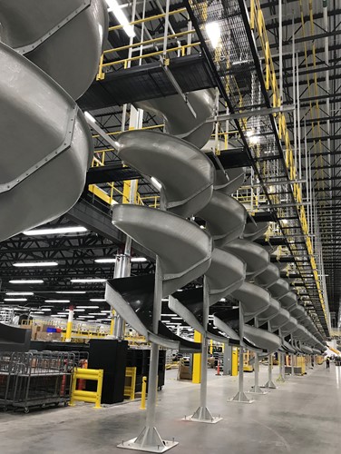 A row of large, silver spiral conveyors extending through a warehouse, supported by steel structures, with a high ceiling and industrial surroundings.