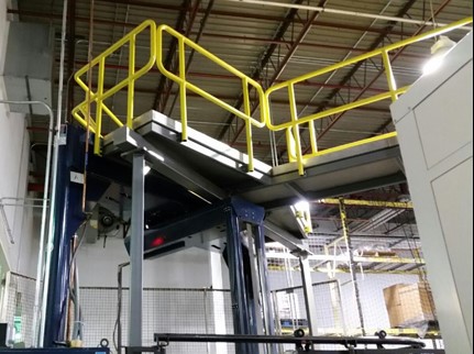 A steel platform with yellow safety handrails, installed inside an industrial facility, supported by sturdy beams and situated near machinery and high ceilings.