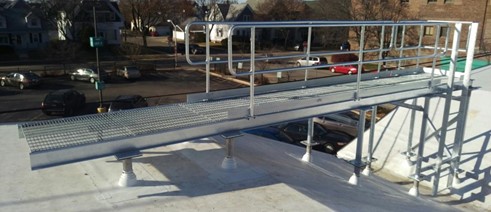 A hot-dipped galvanized steel catwalk with grated platforms and railings, installed on a rooftop. The slightly inclined design provides safe access between levels for maintenance, surrounded by a view of residential and urban streets below.