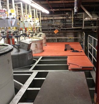 A partially deconstructed industrial platform with exposed structural beams, surrounded by chemical tanks and piping systems. Bright orange safety panels and tools are placed on the remaining sections of the platform, indicating ongoing renovation or demolition work in a manufacturing facility.