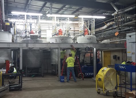 Industrial workers installing a complex chemical processing system at a facility. The setup includes multiple tanks with pipes and valves mounted on a raised platform, surrounded by safety equipment, tools, and a yellow industrial fan for ventilation.