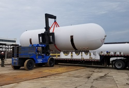A blue Doral forklift is used to lift a large white industrial tank with rigging straps, preparing to load it onto a flatbed truck in an outdoor worksite.
