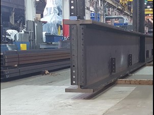 A close-up view of a large steel frame member with bolted connection points, set on the floor of an industrial workspace. Additional steel beams and machinery are visible in the background, emphasizing the manufacturing environment.