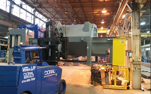 A blue Versa-Lift forklift from Doral Rentals is used to transport a large pass-through blast machine inside a spacious industrial facility with overhead cranes and equipment.