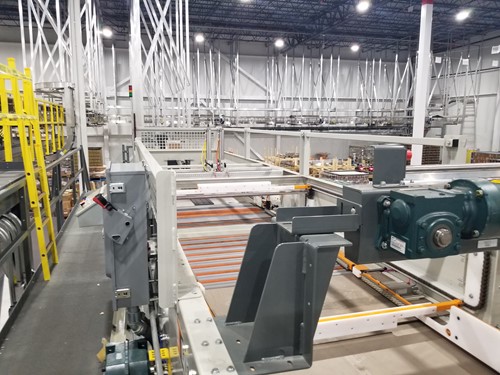 A detailed view of an overhead conveyor system with metal supports and machinery components. The industrial setting features interconnected beams, yellow safety railings, and an organized warehouse environment in the background.