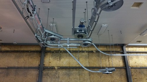 An overhead meat-processing conveyor system viewed from below, displaying suspended metal tracks and pulleys attached to the ceiling in an industrial space with a partially exposed wall.