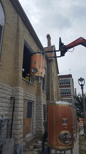 A rigging operation in progress, where a forklift is lifting a large copper brewing tank through an upper window of a brick building, with another tank standing on the ground nearby.