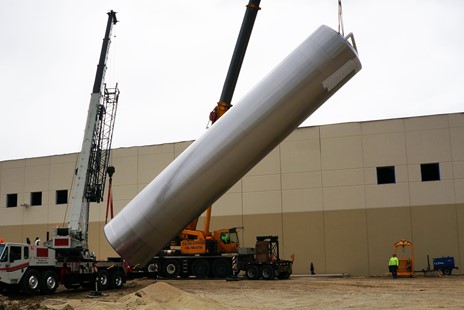 Two cranes are working together to lift and position a large stainless steel silo at an outdoor construction site beside an industrial building, with workers supervising the operation.