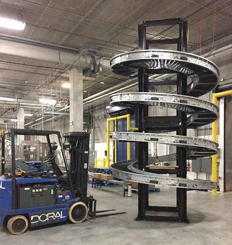 A spiral case conveyor system installed in an industrial facility, featuring a tall, coiled metal track for efficient product movement, with a Doral forklift positioned nearby for support.