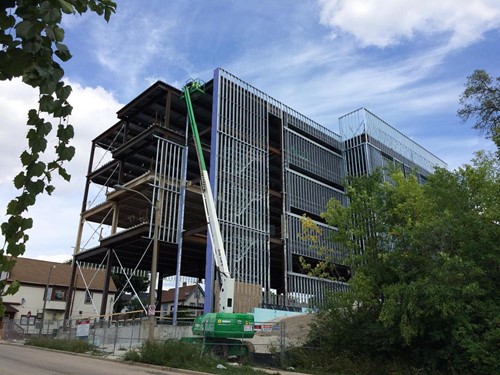 A partially constructed multi-story steel and glass building, with workers using a green lift to install metal framing on the upper levels, surrounded by trees and residential buildings.