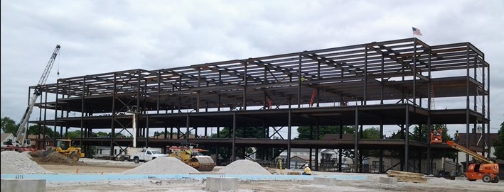 A large multi-level structural steel framework under construction, surrounded by construction equipment and gravel piles, with an American flag atop the structure.