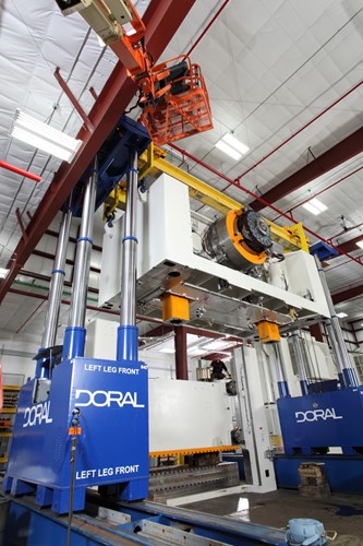 A large industrial setup featuring a blue Doral rigging system being used to stack the crown on a massive 1,375-ton stamping press, with a worker operating from an elevated platform in the background.