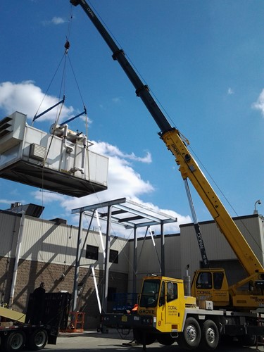 A yellow Doral crane lifts a large industrial unit to place it onto a steel frame structure outside a manufacturing facility, under a bright blue sky with scattered clouds.