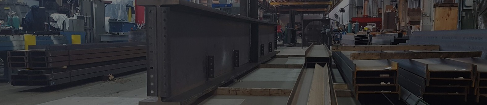 A workshop scene featuring long steel beams and heavy industrial equipment, with workers and machinery visible in the background.
