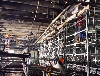 A large industrial space under structural reinforcement, featuring a complex network of scaffolding supporting beams and workers in safety gear performing tasks. Overhead lighting illuminates the work zone, highlighting the extensive temporary steel framework used for stabilization or construction.