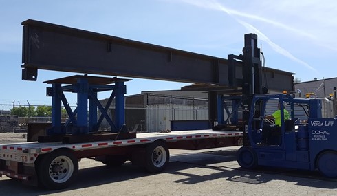 A large steel frame member is being loaded onto a flatbed trailer by a Versa-Lift forklift. The outdoor industrial setting includes the forklift operator ensuring secure transport, with buildings and fences visible in the background.