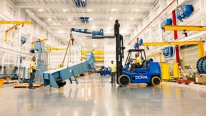Workers operate a blue forklift marked “DORAL” to lift and position a large, angled metal structure in a bright, spacious industrial facility with polished floors, yellow overhead cranes, and neatly organized equipment.