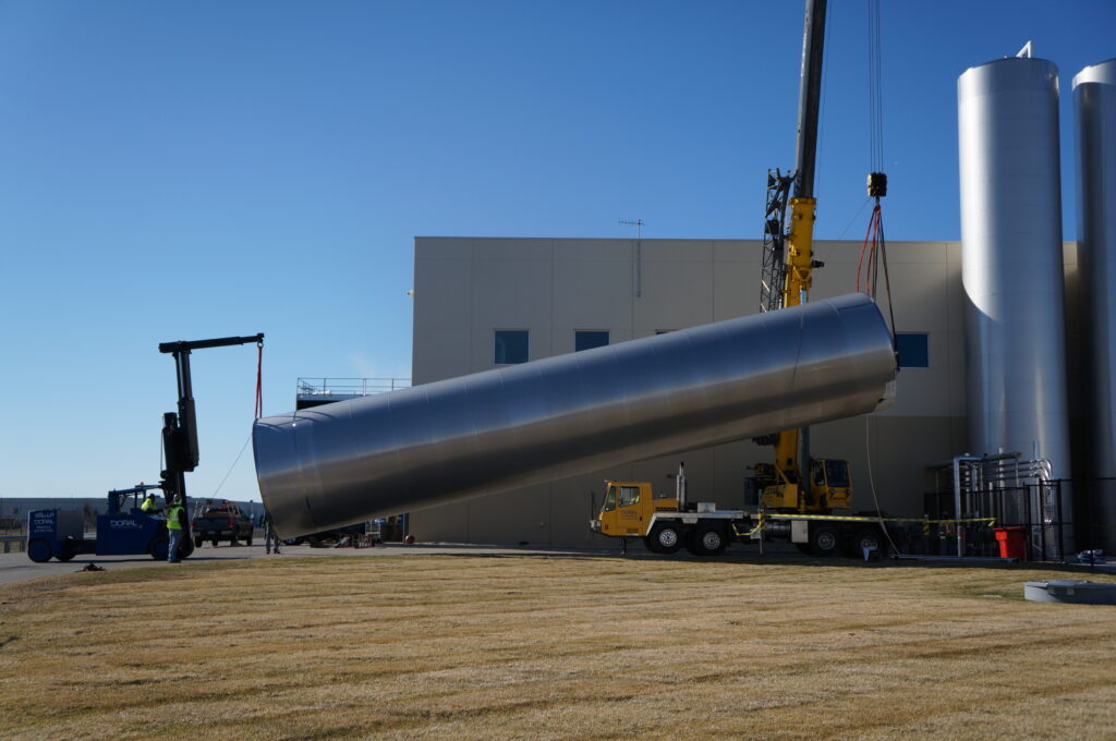 A large stainless steel silo being carefully tilted and positioned upright using a yellow Doral crane and a forklift, set against a modern industrial building on a clear, sunny day.