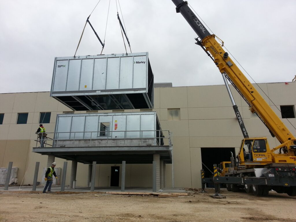 A yellow Doral crane lifting a large SPX Marley industrial unit onto a platform with precision, while workers in safety gear oversee the installation process at a construction site.