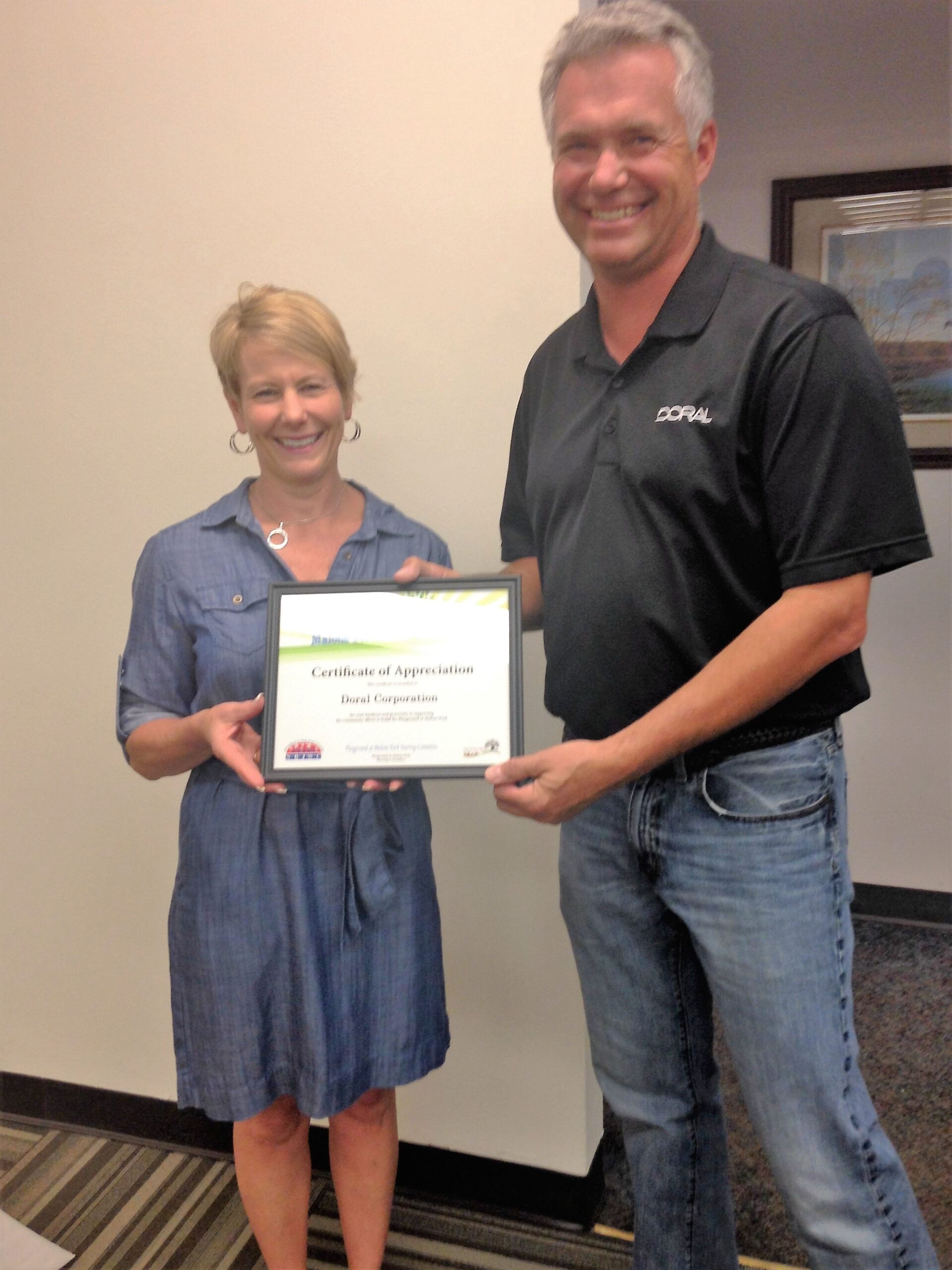 Two smiling individuals pose in an office setting while holding a framed “Certificate of Appreciation” awarded to Doral Corporation. The man wears a black polo shirt with the Doral logo, and the woman wears a denim dress.