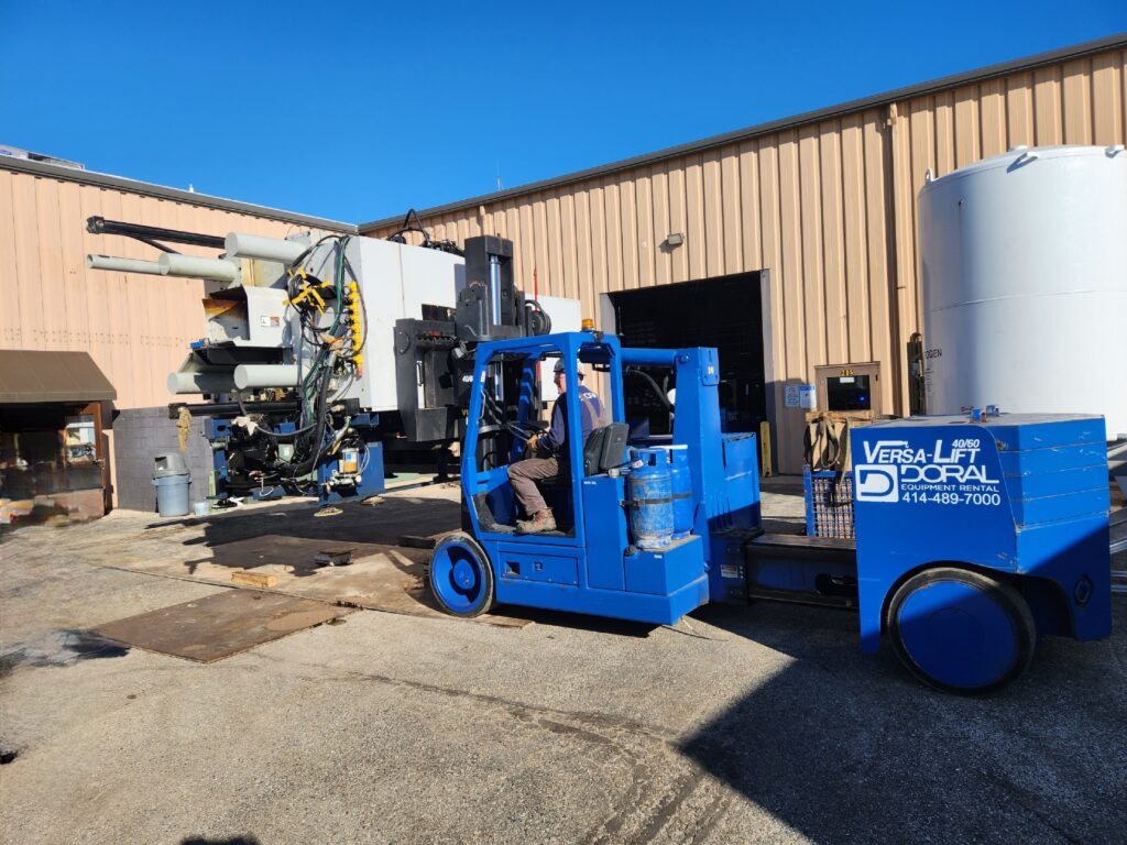 Blue forklift labeled "Versa-Lift Doral" lifts heavy machinery near a beige building in an industrial yard. Operator is visible.