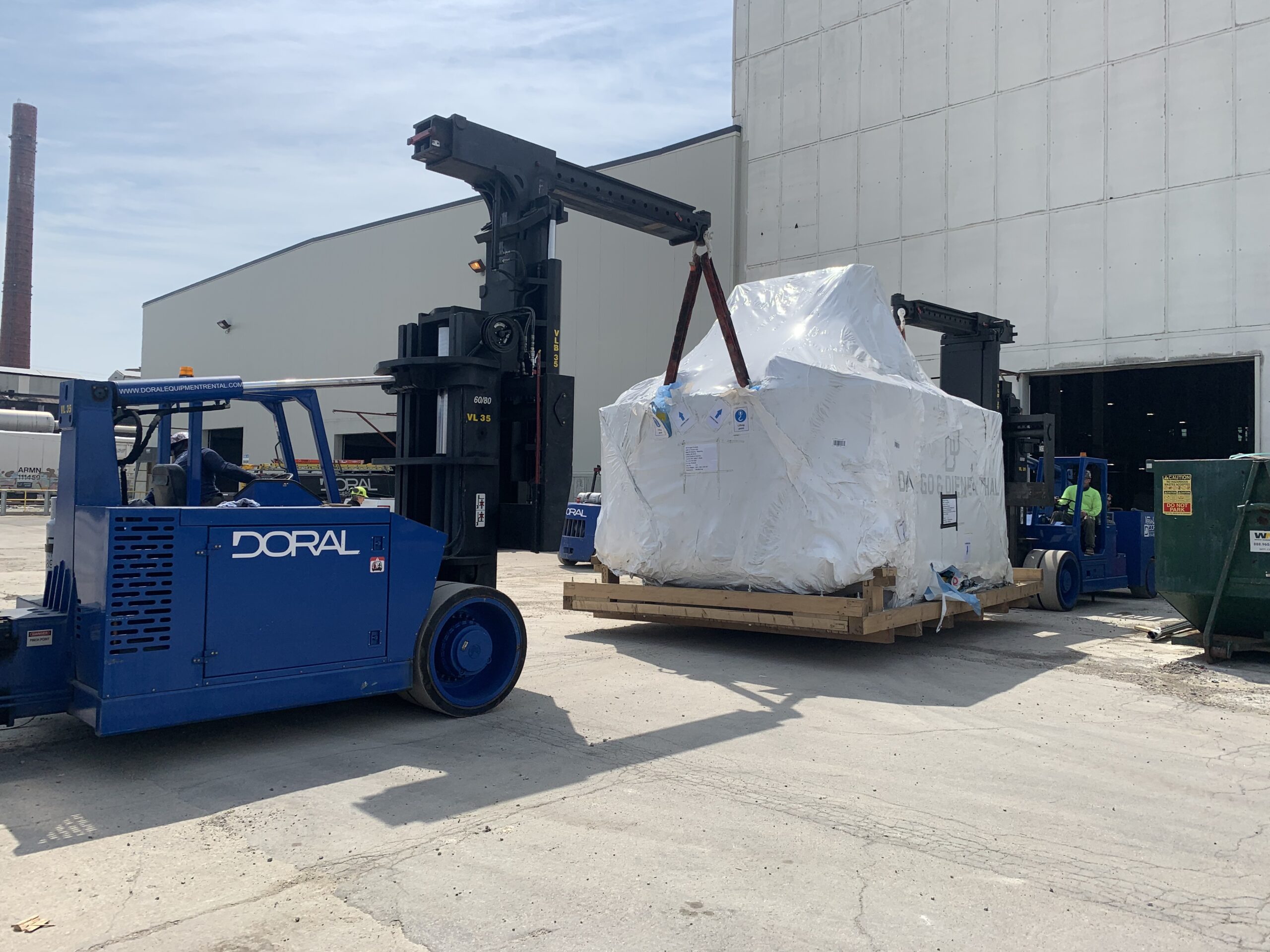 Two blue forklifts are moving a large, white-wrapped object on a wooden pallet outside an industrial building.