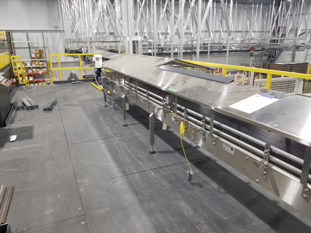 Industrial conveyor belt system in a large warehouse with metal framework, safety railings, and organized storage shelves in the background.