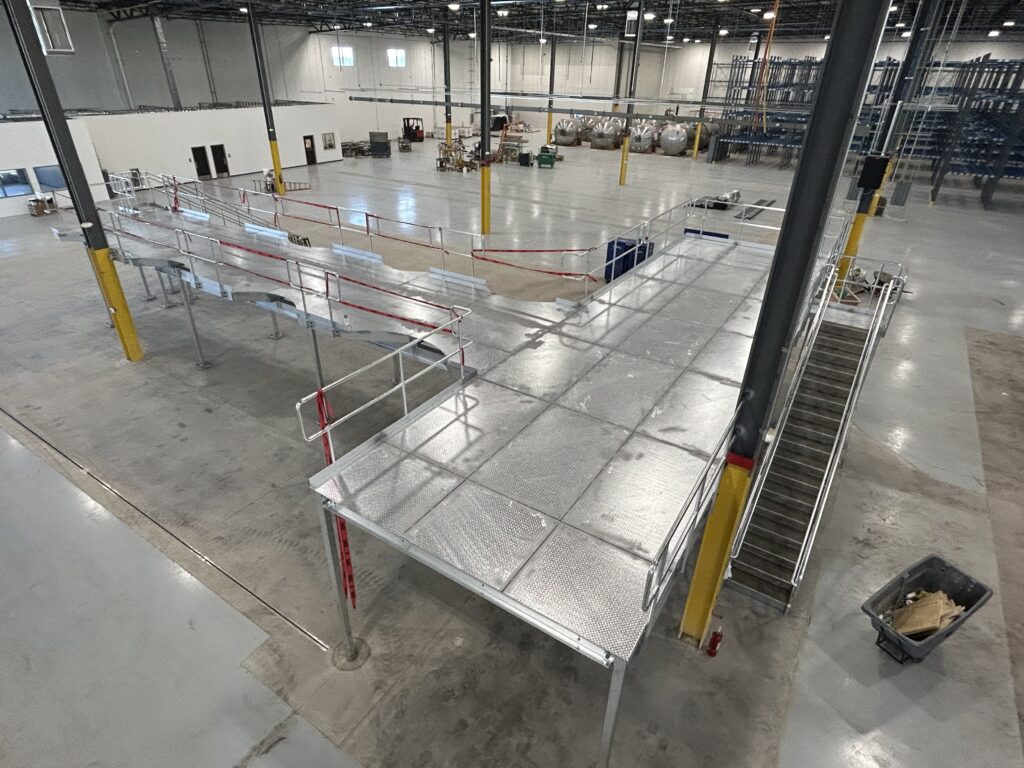 Interior of a large warehouse with metal platforms, yellow pillars, and various equipment scattered around, indicating active setup.
