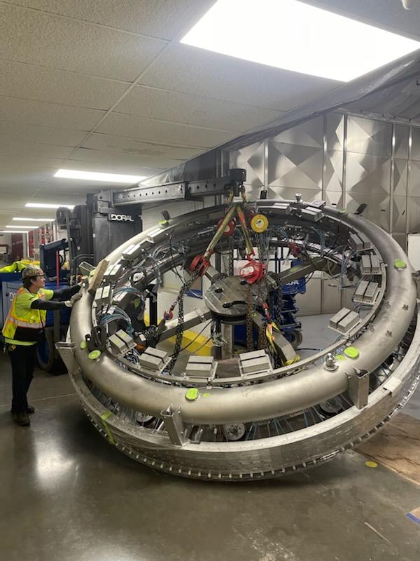 A worker in a safety vest and helmet operates machinery to move a large, circular industrial component in a facility.