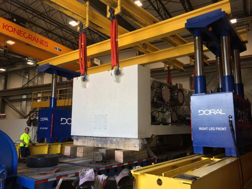 Large industrial machine labeled "DORAL" being lifted by two yellow Konecranes with red straps in a warehouse, workers nearby.