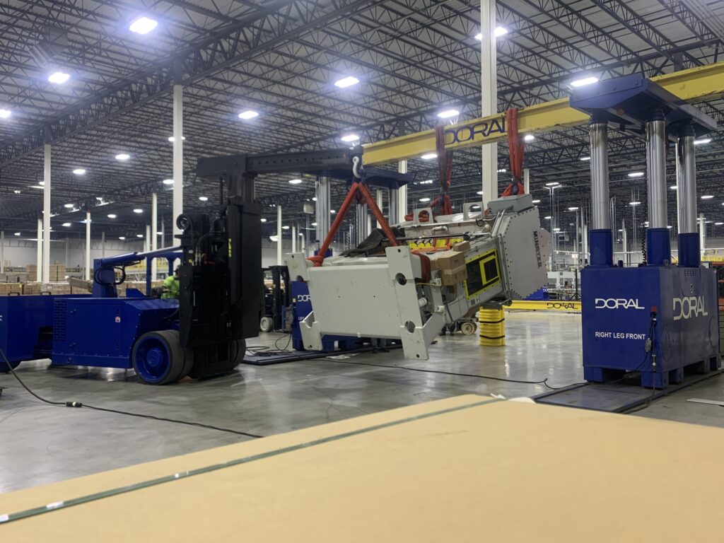 Large industrial warehouse with machinery, including a blue forklift and a yellow crane labeled "Doral," lifting a heavy object.