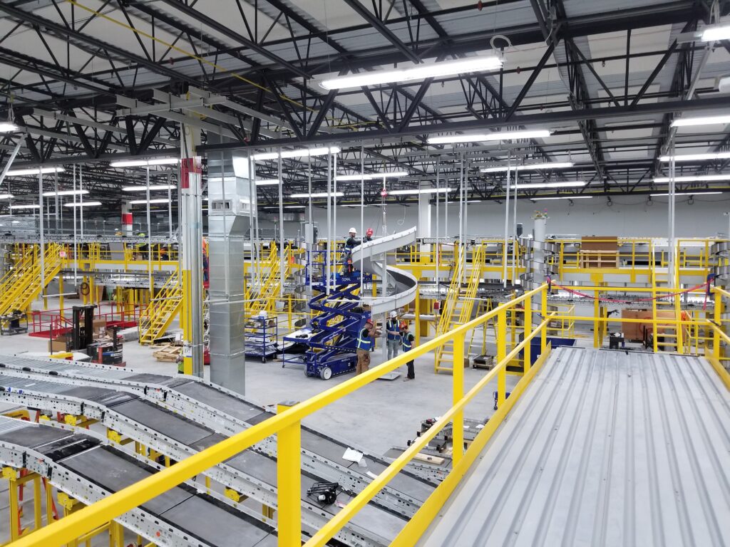 Interior of a busy warehouse with machinery, conveyor belts, safety railings, and workers operating equipment on multiple levels.