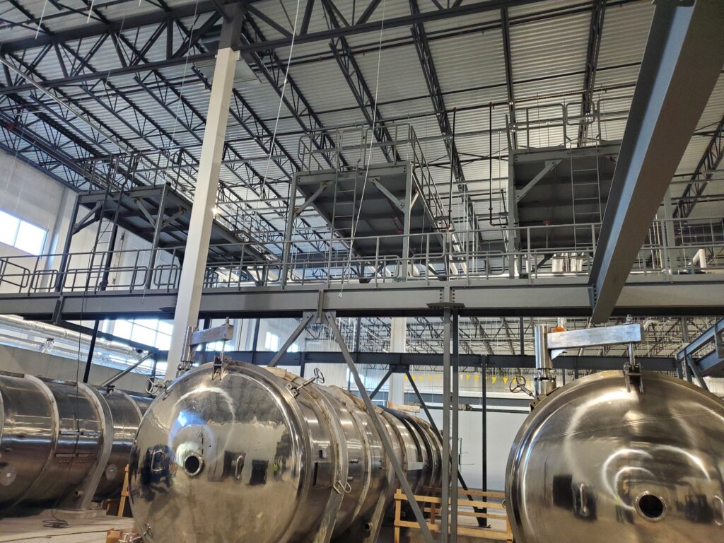 Industrial facility with large cylindrical metal tanks and overhead walkways supported by a steel framework.
