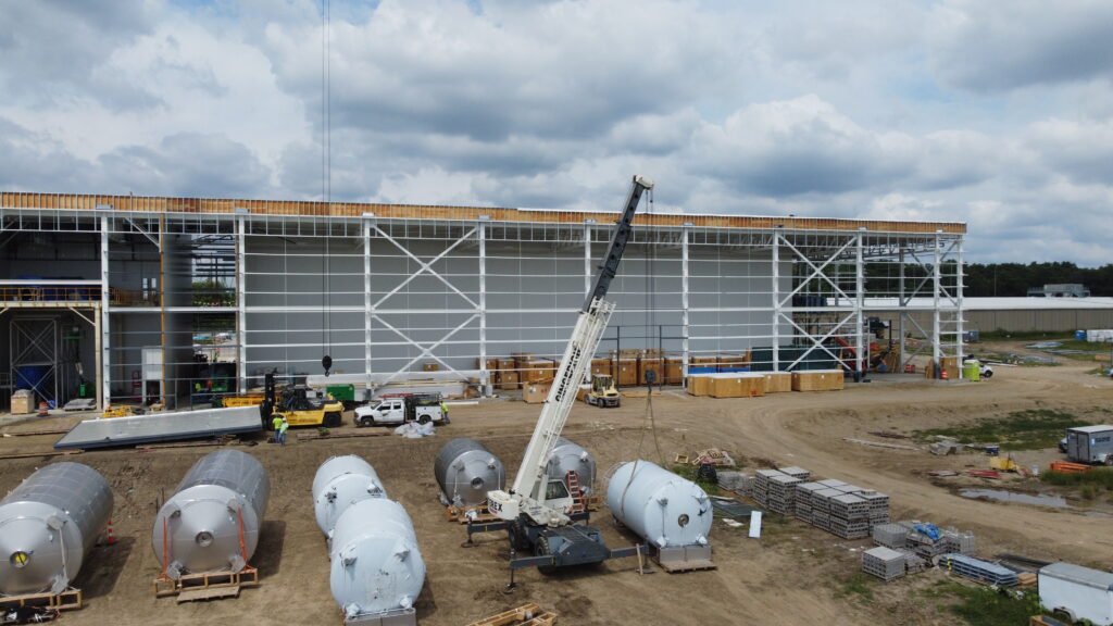 Construction site with a large building, crane lifting materials, and several cylindrical tanks on the ground.
