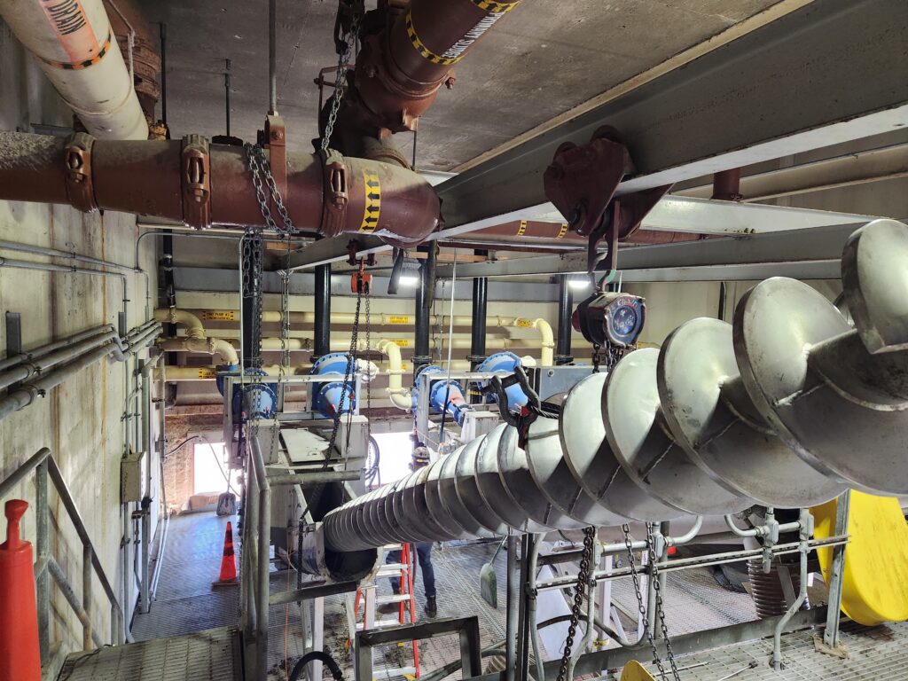Industrial setting with large metal pipes, valves, and machinery. Spiral conveyor in foreground, with safety cones and chains visible.